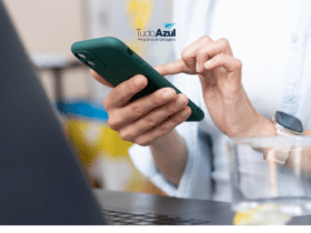Close de uma pessoa segurando um smartphone com capa verde, usando o dispositivo com uma mão enquanto digita com a outra. A pessoa veste uma camisa de cor clara e usa um relógio inteligente com pulseira rosa no pulso esquerdo. Em segundo plano, um laptop está parcialmente visível. No centro superior da imagem, está o logotipo do TudoAzul, Programa de Vantagens da Azul Linhas Aéreas.