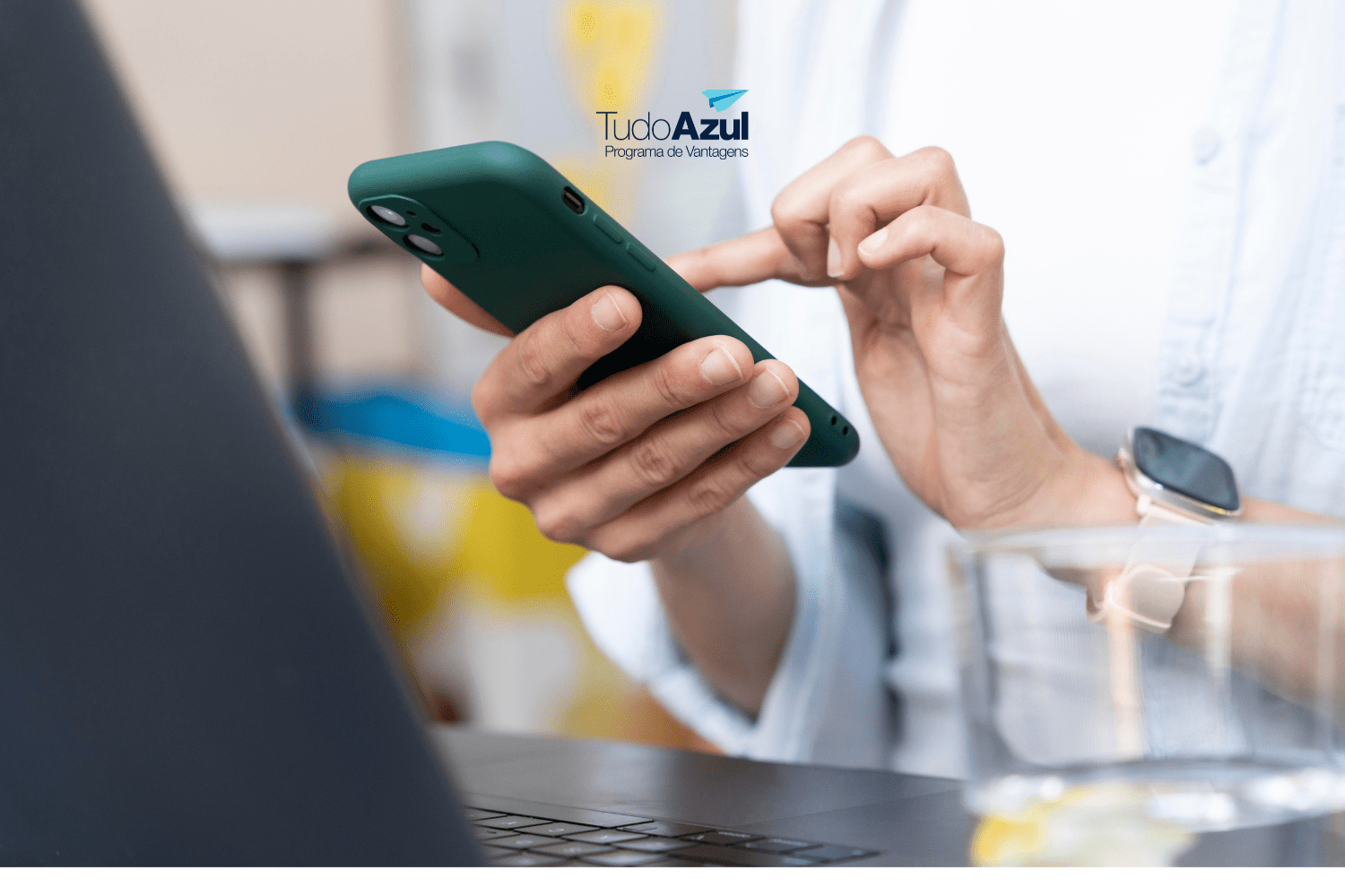 Close de uma pessoa segurando um smartphone com capa verde, usando o dispositivo com uma mão enquanto digita com a outra. A pessoa veste uma camisa de cor clara e usa um relógio inteligente com pulseira rosa no pulso esquerdo. Em segundo plano, um laptop está parcialmente visível. No centro superior da imagem, está o logotipo do TudoAzul, Programa de Vantagens da Azul Linhas Aéreas.