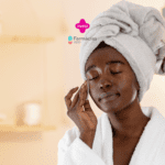 "Uma mulher negra está realizando sua rotina de cuidados com a pele. Ela está de olhos fechados e usando uma toalha enrolada na cabeça e um roupão branco. Ela está passando um algodão no rosto com expressão relaxada e serena. O fundo é suave e desfocado, com uma estante e alguns itens decorativos. No topo da imagem, há os logotipos da Livelo e do Farmácias App.