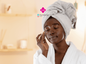 "Uma mulher negra está realizando sua rotina de cuidados com a pele. Ela está de olhos fechados e usando uma toalha enrolada na cabeça e um roupão branco. Ela está passando um algodão no rosto com expressão relaxada e serena. O fundo é suave e desfocado, com uma estante e alguns itens decorativos. No topo da imagem, há os logotipos da Livelo e do Farmácias App.