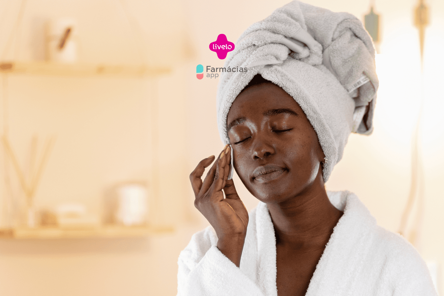 "Uma mulher negra está realizando sua rotina de cuidados com a pele. Ela está de olhos fechados e usando uma toalha enrolada na cabeça e um roupão branco. Ela está passando um algodão no rosto com expressão relaxada e serena. O fundo é suave e desfocado, com uma estante e alguns itens decorativos. No topo da imagem, há os logotipos da Livelo e do Farmácias App.