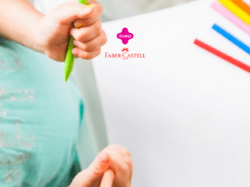 Mãos de uma criança segurando lápis de cor verde e laranja, enquanto desenha em um papel branco. No canto direito da imagem, há vários lápis de cor em diferentes cores (rosa, amarelo, azul, vermelho e laranja) alinhados sobre a mesa. A criança está vestindo uma camiseta verde clara com detalhes florais. No centro superior da imagem, estão os logotipos da Livelo e Faber-Castell, indicando uma parceria entre as duas marcas. A imagem transmite uma sensação de criatividade e diversão.