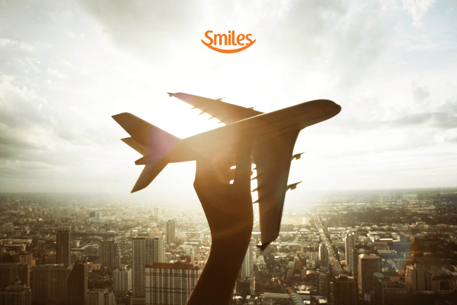 A imagem mostra a silhueta de um avião sendo segurado por uma mão contra um fundo de céu claro e ensolarado. A cidade aparece embaixo, com vários prédios e construções visíveis, mas desfocados. O logotipo da Smiles, em laranja, está localizado no topo central da imagem. A composição transmite uma sensação de voo e liberdade.