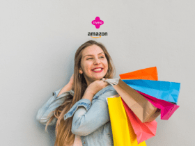 Na imagem, vemos uma mulher jovem e sorridente segurando várias sacolas de compras coloridas. Ela está vestida com uma jaqueta jeans e parece estar muito feliz com suas compras. No topo da imagem, há os logotipos da Livelo e da Amazon, indicando uma possível parceria ou promoção entre as duas empresas. A mulher está em pé contra uma parede cinza clara, que serve como fundo simples para destacar seu sorriso e as sacolas de compras vibrantes.