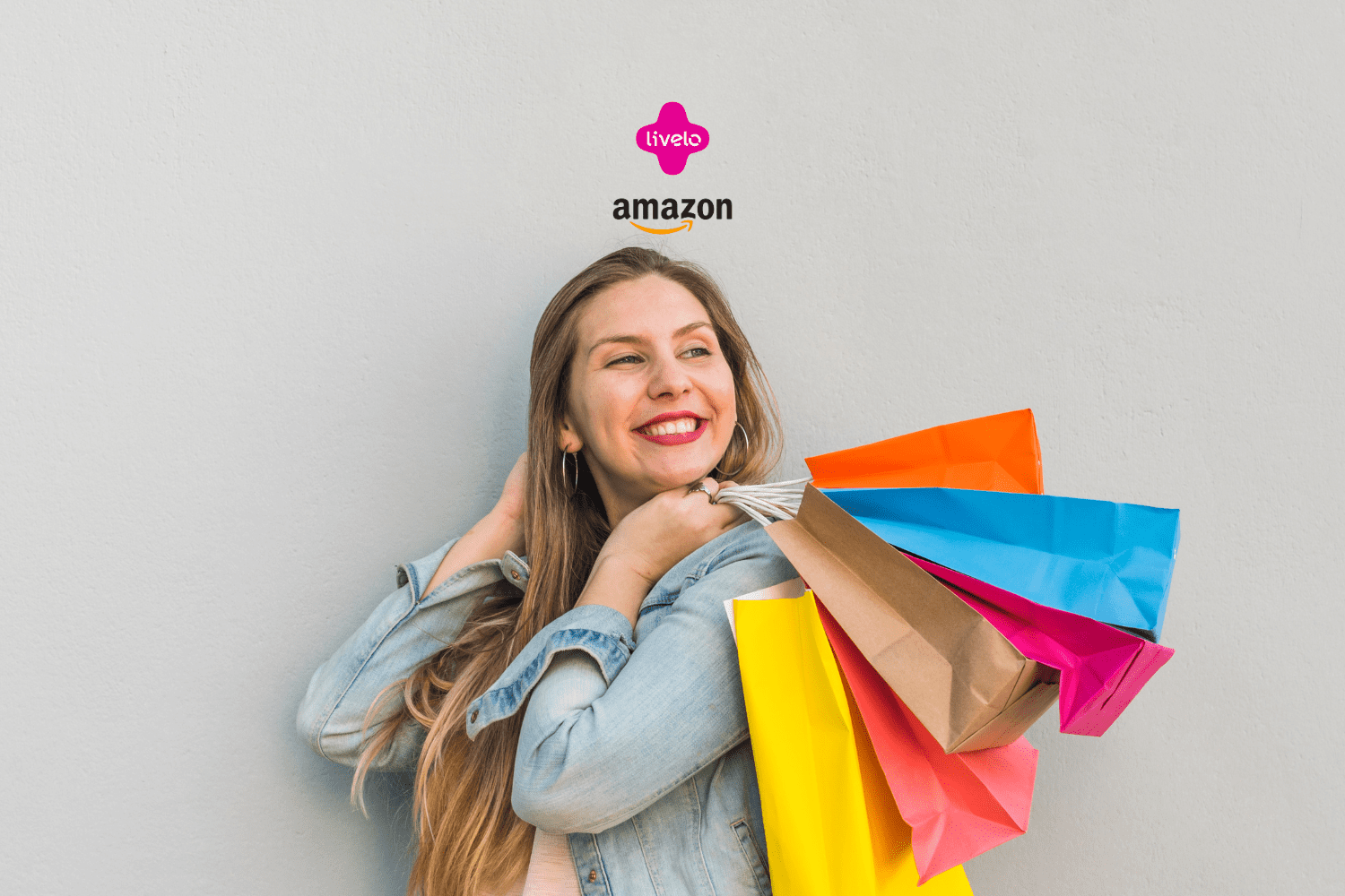 Na imagem, vemos uma mulher jovem e sorridente segurando várias sacolas de compras coloridas. Ela está vestida com uma jaqueta jeans e parece estar muito feliz com suas compras. No topo da imagem, há os logotipos da Livelo e da Amazon, indicando uma possível parceria ou promoção entre as duas empresas. A mulher está em pé contra uma parede cinza clara, que serve como fundo simples para destacar seu sorriso e as sacolas de compras vibrantes.