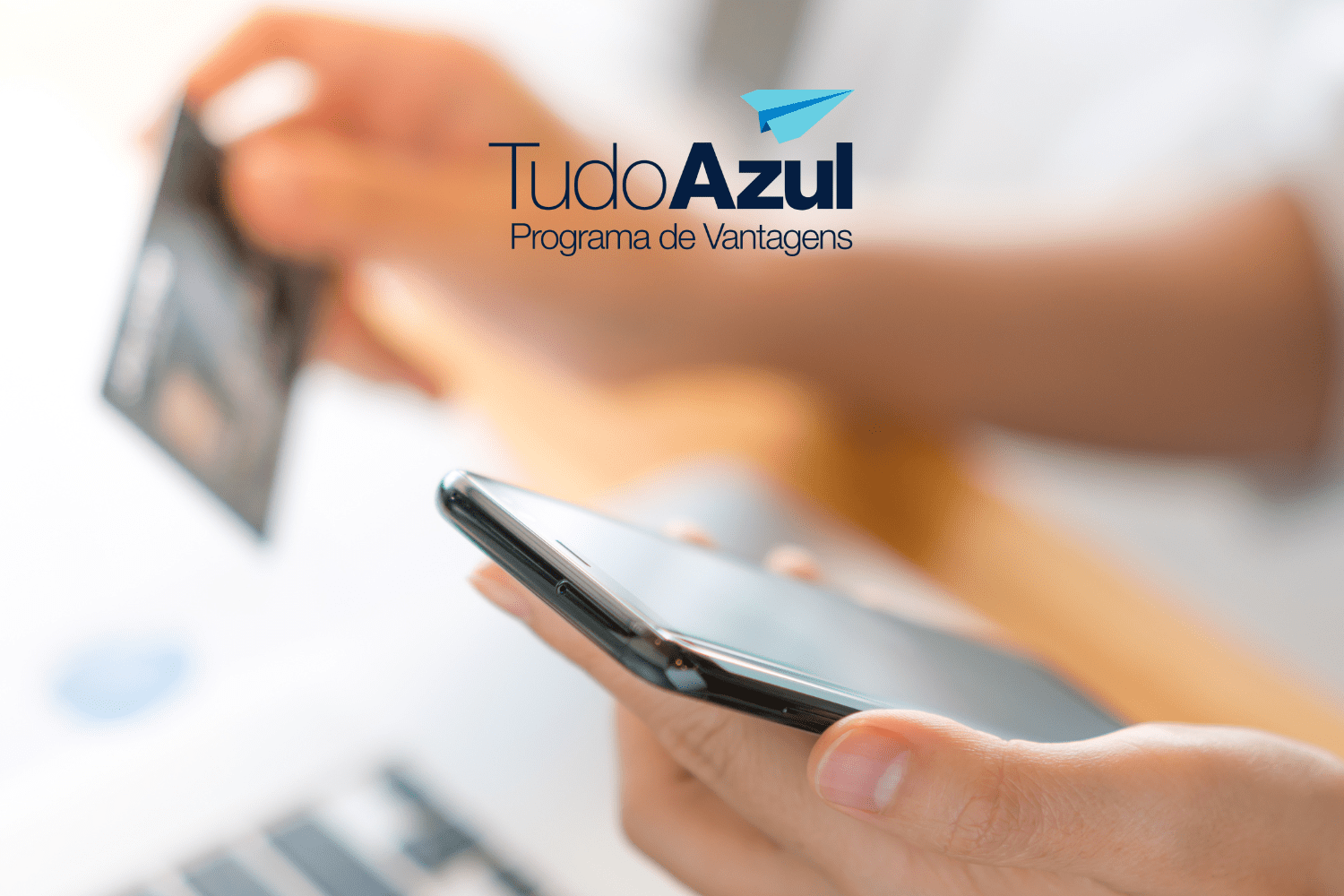 A imagem mostra uma pessoa segurando um smartphone com uma mão e um cartão de crédito com a outra. A imagem está levemente desfocada ao fundo, focando nas mãos e nos objetos que estão sendo segurados. No canto superior direito da imagem, há o logotipo do programa de vantagens "TudoAzul", com um ícone de avião de papel azul.