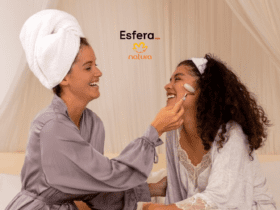 A imagem mostra duas mulheres sorrindo e interagindo em um ambiente acolhedor, com cortinas claras ao fundo. A mulher à esquerda está com uma toalha branca enrolada na cabeça e veste um robe cinza, enquanto segura um massageador facial de quartzo rosa no rosto da outra mulher. A mulher à direita, com cabelos cacheados soltos e uma faixa branca na cabeça, veste uma camisola branca com detalhes de renda e um robe leve. Ambas parecem estar se divertindo em um momento de cuidados pessoais. Acima das mulheres, estão os logotipos da Esfera e da Natura, centralizados na parte superior da imagem.