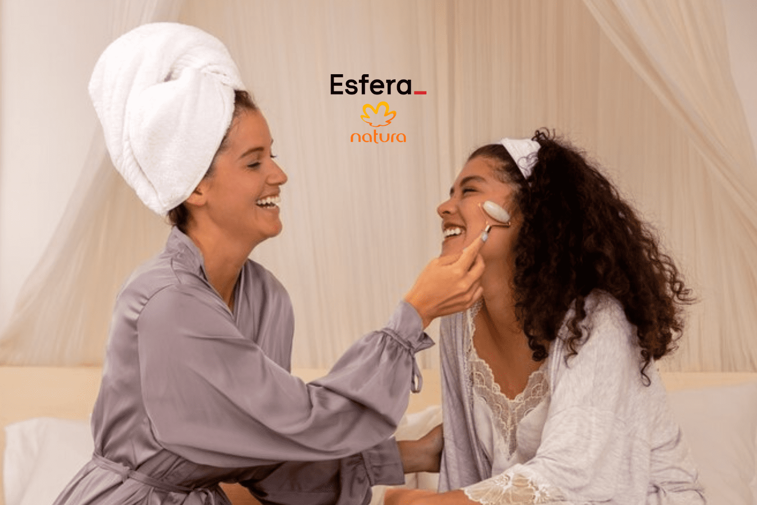 A imagem mostra duas mulheres sorrindo e interagindo em um ambiente acolhedor, com cortinas claras ao fundo. A mulher à esquerda está com uma toalha branca enrolada na cabeça e veste um robe cinza, enquanto segura um massageador facial de quartzo rosa no rosto da outra mulher. A mulher à direita, com cabelos cacheados soltos e uma faixa branca na cabeça, veste uma camisola branca com detalhes de renda e um robe leve. Ambas parecem estar se divertindo em um momento de cuidados pessoais. Acima das mulheres, estão os logotipos da Esfera e da Natura, centralizados na parte superior da imagem.