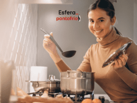 Uma mulher jovem e sorridente está cozinhando em uma cozinha moderna. Ela segura uma concha com uma mão e uma tampa de panela com a outra, enquanto está de frente para um fogão com uma panela de aço inoxidável. Ela veste uma blusa de gola alta bege. Ao fundo, vemos uma parede com revestimento decorativo e utensílios de cozinha. No canto superior direito da imagem, está o logo da Esfera e Pontofrio.