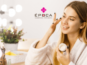 "Uma mulher jovem, com cabelo loiro comprido e usando um roupão branco felpudo, sorri enquanto aplica creme no rosto. Ela segura um pote de creme na mão esquerda e usa a mão direita para aplicar o produto. Ao fundo, luzes brilhantes e uma planta decorativa. Ao lado dela, pincéis de maquiagem em um suporte dourado. Logotipo da Livelo e nome da loja Época Cosméticos Perfumaria visíveis no canto superior esquerdo."