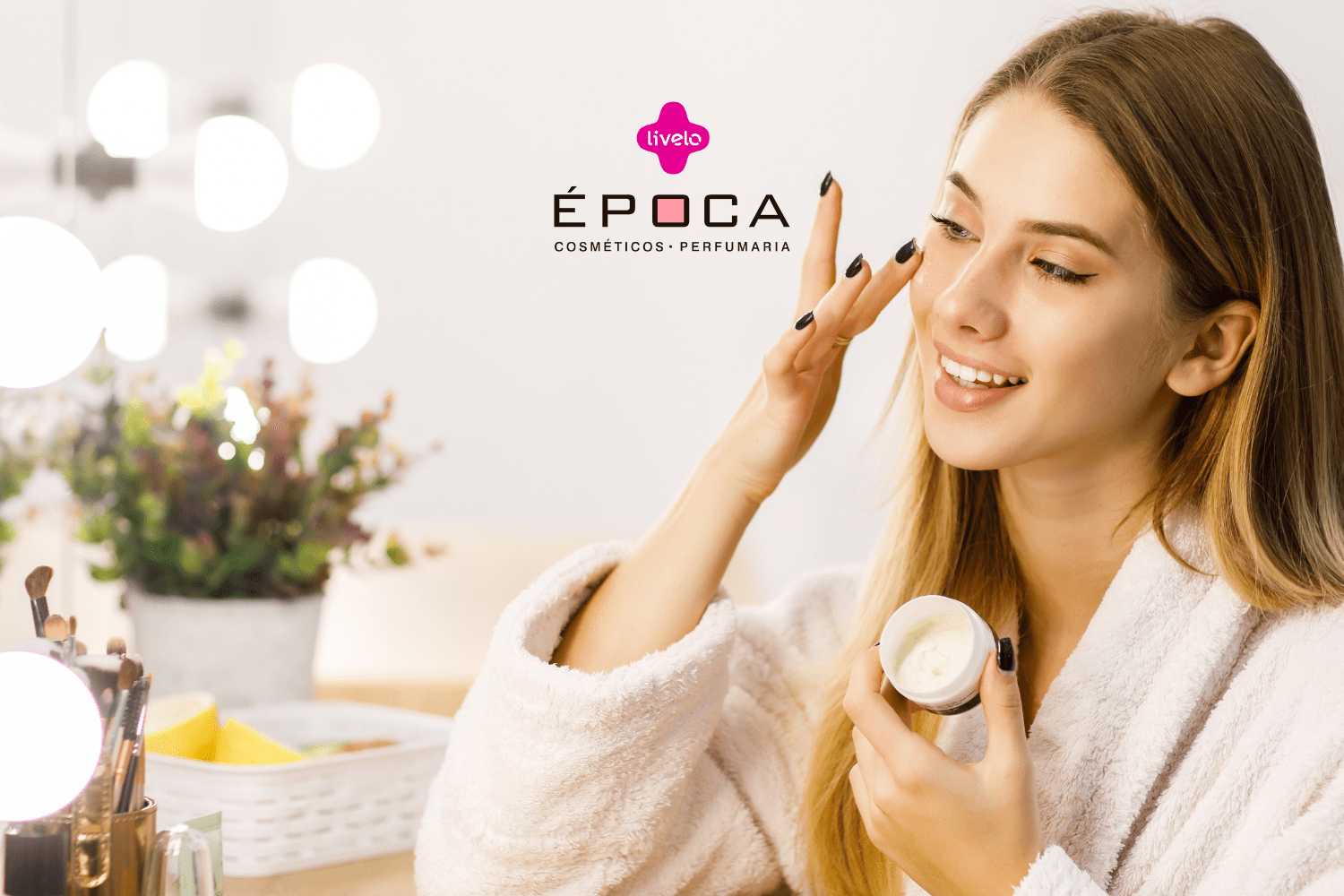 "Uma mulher jovem, com cabelo loiro comprido e usando um roupão branco felpudo, sorri enquanto aplica creme no rosto. Ela segura um pote de creme na mão esquerda e usa a mão direita para aplicar o produto. Ao fundo, luzes brilhantes e uma planta decorativa. Ao lado dela, pincéis de maquiagem em um suporte dourado. Logotipo da Livelo e nome da loja Época Cosméticos Perfumaria visíveis no canto superior esquerdo."