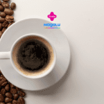 A imagem mostra uma xícara de café expresso em um pires branco, vista de cima, posicionada à direita de um fundo claro. A xícara está cheia de café preto, com uma fina camada de espuma marrom clara na superfície. À esquerda, espalhados sobre a mesa, estão diversos grãos de café marrons, que criam um contraste com a superfície branca e adicionam textura à composição. Acima da xícara, no centro da imagem, estão os logotipos da Livelo e da loja Magalu.