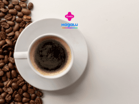 A imagem mostra uma xícara de café expresso em um pires branco, vista de cima, posicionada à direita de um fundo claro. A xícara está cheia de café preto, com uma fina camada de espuma marrom clara na superfície. À esquerda, espalhados sobre a mesa, estão diversos grãos de café marrons, que criam um contraste com a superfície branca e adicionam textura à composição. Acima da xícara, no centro da imagem, estão os logotipos da Livelo e da loja Magalu.