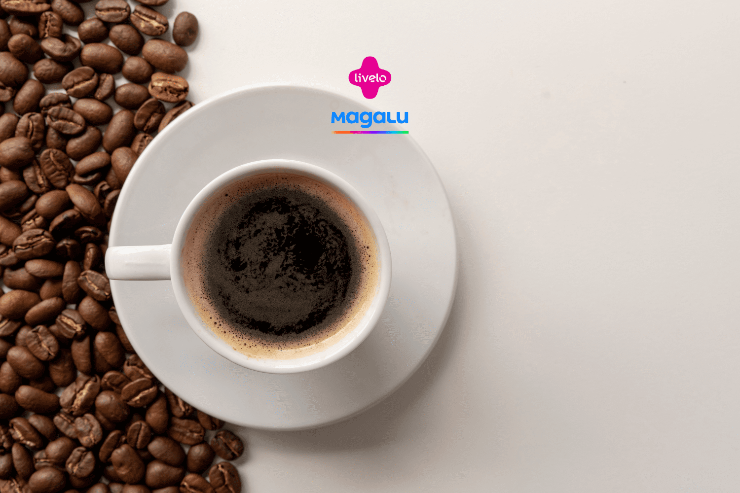 A imagem mostra uma xícara de café expresso em um pires branco, vista de cima, posicionada à direita de um fundo claro. A xícara está cheia de café preto, com uma fina camada de espuma marrom clara na superfície. À esquerda, espalhados sobre a mesa, estão diversos grãos de café marrons, que criam um contraste com a superfície branca e adicionam textura à composição. Acima da xícara, no centro da imagem, estão os logotipos da Livelo e da loja Magalu.