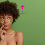 Uma mulher com cabelos cacheados e expressão pensativa está de pé contra um fundo verde. Ela tem dois adesivos verdes em forma de meia-lua sob os olhos, usados para cuidados com a pele. Acima dela, estão os logotipos da Livelo e da Eudora em rosa e roxo, respectivamente.