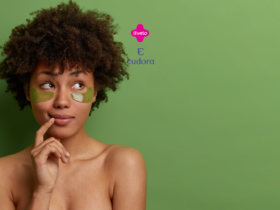 Uma mulher com cabelos cacheados e expressão pensativa está de pé contra um fundo verde. Ela tem dois adesivos verdes em forma de meia-lua sob os olhos, usados para cuidados com a pele. Acima dela, estão os logotipos da Livelo e da Eudora em rosa e roxo, respectivamente.