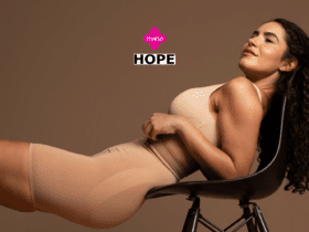 Uma mulher de pele morena e cabelos cacheados, vestida com um conjunto de roupas íntimas de cor nude, está reclinada em uma cadeira preta. Ela parece relaxada e confiante, olhando para cima, com uma expressão serena em seu rosto. Ao fundo, há um tom marrom uniforme. Acima de sua cabeça, estão os logotipos das marcas 'Livelo' em rosa e 'Hope' em preto e branco