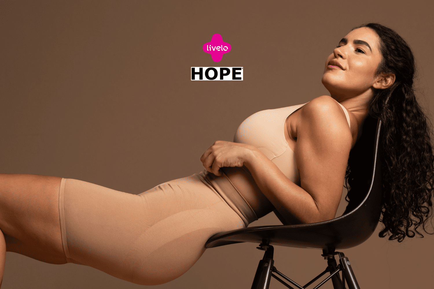 Uma mulher de pele morena e cabelos cacheados, vestida com um conjunto de roupas íntimas de cor nude, está reclinada em uma cadeira preta. Ela parece relaxada e confiante, olhando para cima, com uma expressão serena em seu rosto. Ao fundo, há um tom marrom uniforme. Acima de sua cabeça, estão os logotipos das marcas 'Livelo' em rosa e 'Hope' em preto e branco