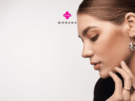 "Uma mulher com cabelo castanho claro, preso em um coque elegante, está de perfil contra um fundo branco. Ela usa um brinco dourado com um design geométrico em forma de losango, adornado com padrões em preto e dourado. Na mão, ela exibe um anel dourado que complementa o brinco. A expressão no rosto dela é serena e concentrada. Acima, estão os logotipos das marcas 'Livelo' em rosa e 'Morana' em preto".
