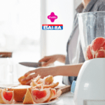 Uma pessoa está em uma cozinha iluminada, preparando frutas frescas. Na imagem, vemos um liquidificador transparente contendo pedaços de grapefruit rosa prontos para serem batidos. Ao fundo, a pessoa segura uma faca enquanto corta as frutas em uma tábua de madeira, com uma seleção de grapefruit já cortada em fatias repousando em um prato ao lado. Acima da cena, estão os logotipos das marcas Livelo e Casas Bahia, sugerindo uma colaboração ou promoção conjunta.