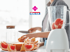 Uma pessoa está em uma cozinha iluminada, preparando frutas frescas. Na imagem, vemos um liquidificador transparente contendo pedaços de grapefruit rosa prontos para serem batidos. Ao fundo, a pessoa segura uma faca enquanto corta as frutas em uma tábua de madeira, com uma seleção de grapefruit já cortada em fatias repousando em um prato ao lado. Acima da cena, estão os logotipos das marcas Livelo e Casas Bahia, sugerindo uma colaboração ou promoção conjunta.