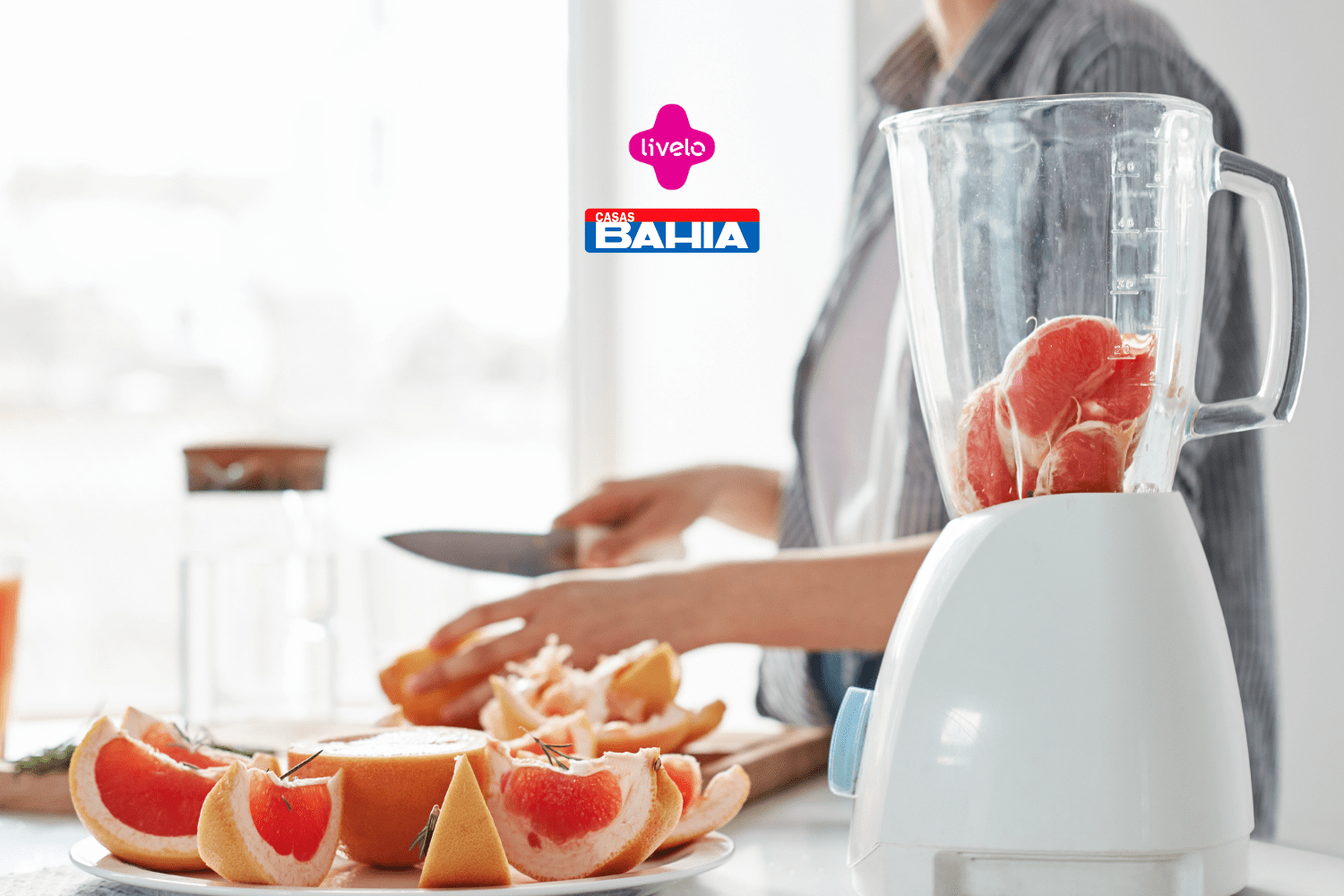 Uma pessoa está em uma cozinha iluminada, preparando frutas frescas. Na imagem, vemos um liquidificador transparente contendo pedaços de grapefruit rosa prontos para serem batidos. Ao fundo, a pessoa segura uma faca enquanto corta as frutas em uma tábua de madeira, com uma seleção de grapefruit já cortada em fatias repousando em um prato ao lado. Acima da cena, estão os logotipos das marcas Livelo e Casas Bahia, sugerindo uma colaboração ou promoção conjunta.