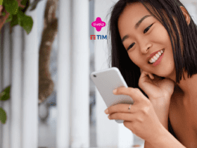 Uma jovem mulher asiática, com cabelo preto e curto, está sorrindo enquanto segura um smartphone em uma das mãos. Ela está sentada em um ambiente externo, cercada por colunas brancas e vegetação ao fundo. A mulher parece estar relaxada e envolvida em algo interessante no telefone. Acima dela, estão os logotipos das marcas Livelo e TIM, sugerindo uma parceria ou promoção associada à tecnologia e comunicação.