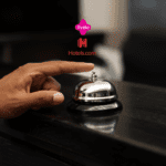 Mão de uma pessoa prestes a tocar uma campainha de balcão em uma recepção de hotel. Acima da campainha, estão os logos da Livelo e do Hotels.com, indicando uma parceria relacionada a reservas de hospedagem.