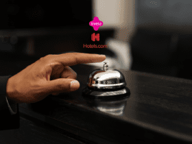 Mão de uma pessoa prestes a tocar uma campainha de balcão em uma recepção de hotel. Acima da campainha, estão os logos da Livelo e do Hotels.com, indicando uma parceria relacionada a reservas de hospedagem.
