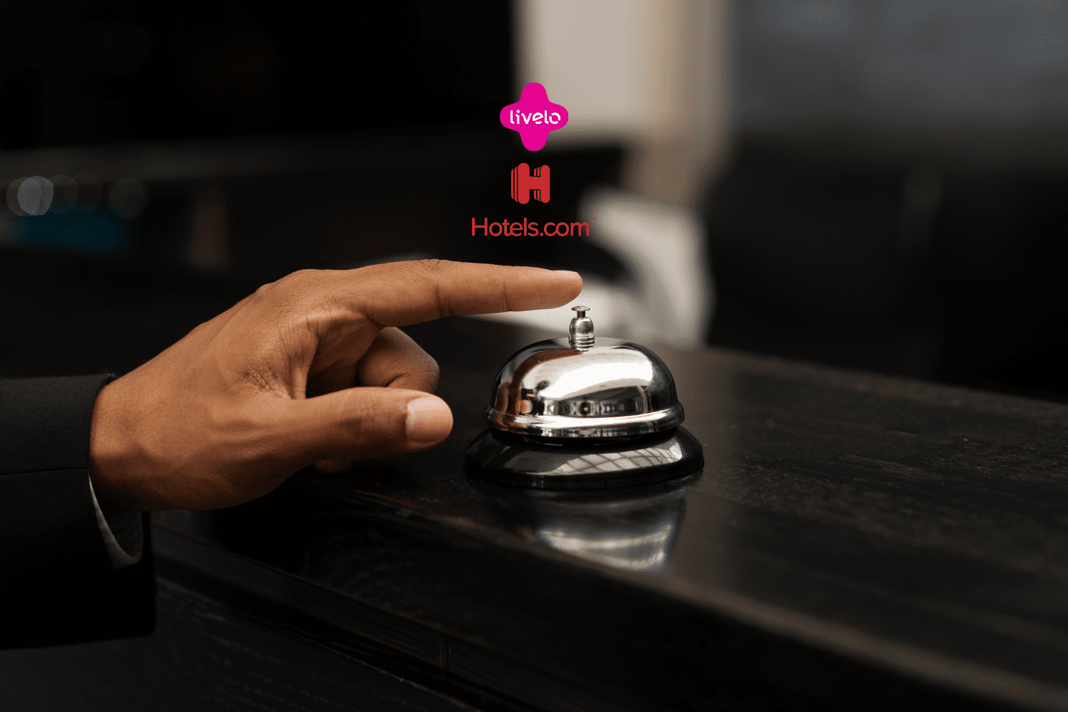 Mão de uma pessoa prestes a tocar uma campainha de balcão em uma recepção de hotel. Acima da campainha, estão os logos da Livelo e do Hotels.com, indicando uma parceria relacionada a reservas de hospedagem.