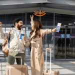 Casal sorridente em frente a um aeroporto, segurando passagens aéreas e levantando os braços em comemoração. Eles têm malas de viagem ao lado. Acima deles está o logo da Smiles, indicando uma viagem feliz e recompensadora.