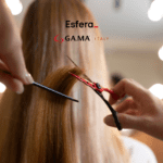 Cabeleireiro cortando o cabelo liso e loiro de uma pessoa com uma tesoura vermelha e um pente, em um salão de beleza. Acima, estão os logos da Esfera e da GA.MA Italy, indicando uma parceria relacionada a cuidados com os cabelos.