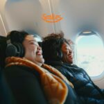 Duas mulheres sentadas em um avião, usando fones de ouvido, estão sorrindo enquanto apreciam o voo. Através da janela, o céu está azul com nuvens brancas. Acima delas, está o logo laranja da Smiles, transmitindo uma sensação de alegria e relaxamento durante a viagem.