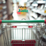 Carrinho de supermercado vazio, visto de trás, em um corredor de supermercado desfocado. Acima do carrinho, estão os logos da Esfera e do Justo, sugerindo uma parceria relacionada a compras e supermercados.