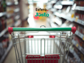 Carrinho de supermercado vazio, visto de trás, em um corredor de supermercado desfocado. Acima do carrinho, estão os logos da Esfera e do Justo, sugerindo uma parceria relacionada a compras e supermercados.