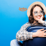 Mulher jovem, sorridente, usando óculos e chapéu de palha, abraçada a uma mala de viagem azul, contra um fundo azul. Acima dela, está o logo da Smiles, sugerindo entusiasmo por viagens.