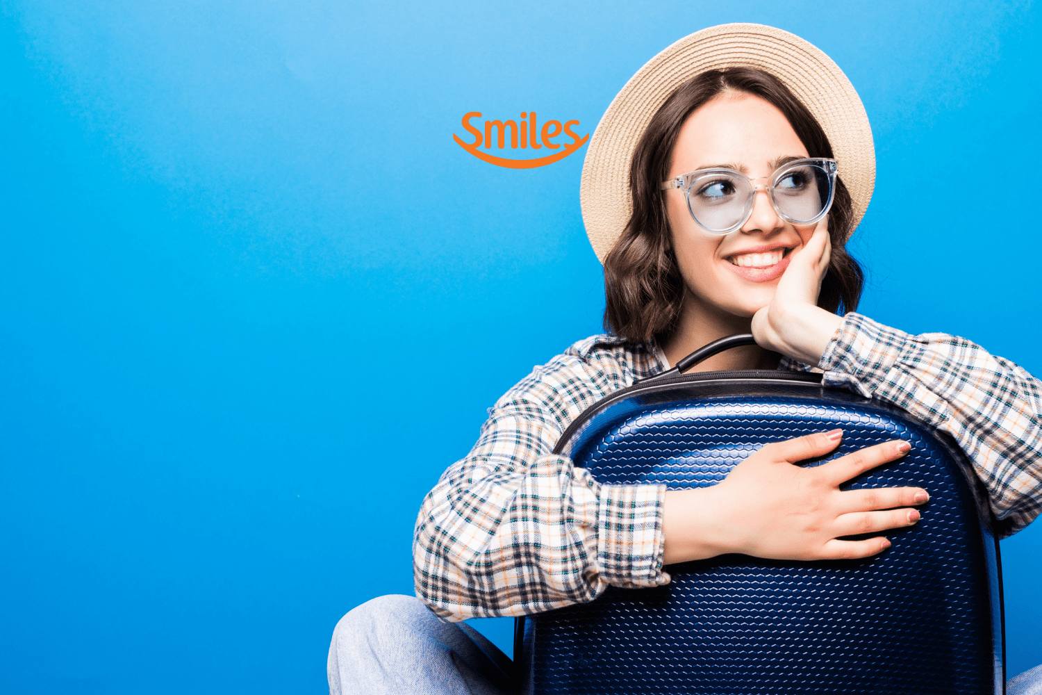 Mulher jovem, sorridente, usando óculos e chapéu de palha, abraçada a uma mala de viagem azul, contra um fundo azul. Acima dela, está o logo da Smiles, sugerindo entusiasmo por viagens.