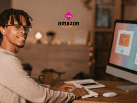 Homem negro com cabelo trançado e óculos, sorrindo para a câmera enquanto trabalha em um computador de mesa em um ambiente aconchegante. No canto superior, os logos da Livelo e da Amazon aparecem juntos.