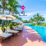 Piscina em um resort cercada por espreguiçadeiras com guarda-sóis brancos, palmeiras e um céu azul claro. Acima da cena, estão os logos da Livelo e do Beach Park, sugerindo um ambiente de lazer e relaxamento em um destino tropical.