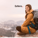 Mulher sentada em uma rocha no alto de uma montanha, sorrindo, com uma mochila de trilha nas costas. Ao fundo, uma vista panorâmica de uma cidade envolta em neblina. Acima dela, está o logo da Esfera, sugerindo uma aventura ao ar livre.