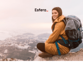 Mulher sentada em uma rocha no alto de uma montanha, sorrindo, com uma mochila de trilha nas costas. Ao fundo, uma vista panorâmica de uma cidade envolta em neblina. Acima dela, está o logo da Esfera, sugerindo uma aventura ao ar livre.