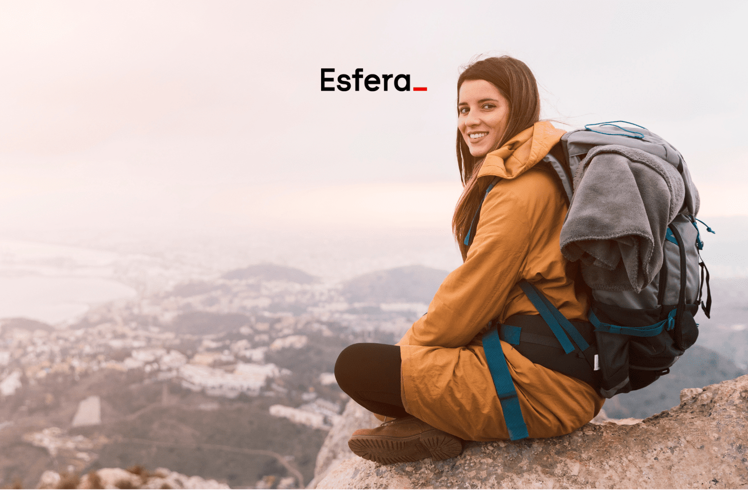 Mulher sentada em uma rocha no alto de uma montanha, sorrindo, com uma mochila de trilha nas costas. Ao fundo, uma vista panorâmica de uma cidade envolta em neblina. Acima dela, está o logo da Esfera, sugerindo uma aventura ao ar livre.