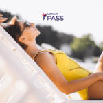 Mulher deitada em uma espreguiçadeira na praia, usando maiô amarelo e óculos de sol, relaxando sob o sol. Acima dela, está o logo do LATAM Pass, sugerindo uma experiência de viagem tranquila e relaxante