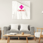 Sala de estar decorada com sofá cinza, almofadas brancas e mesa de centro de madeira. Uma luminária de chão branca e uma poltrona com estrutura de madeira complementam o ambiente. Na parede, um quadro com a logomarca da Livelo e da Camicado.
