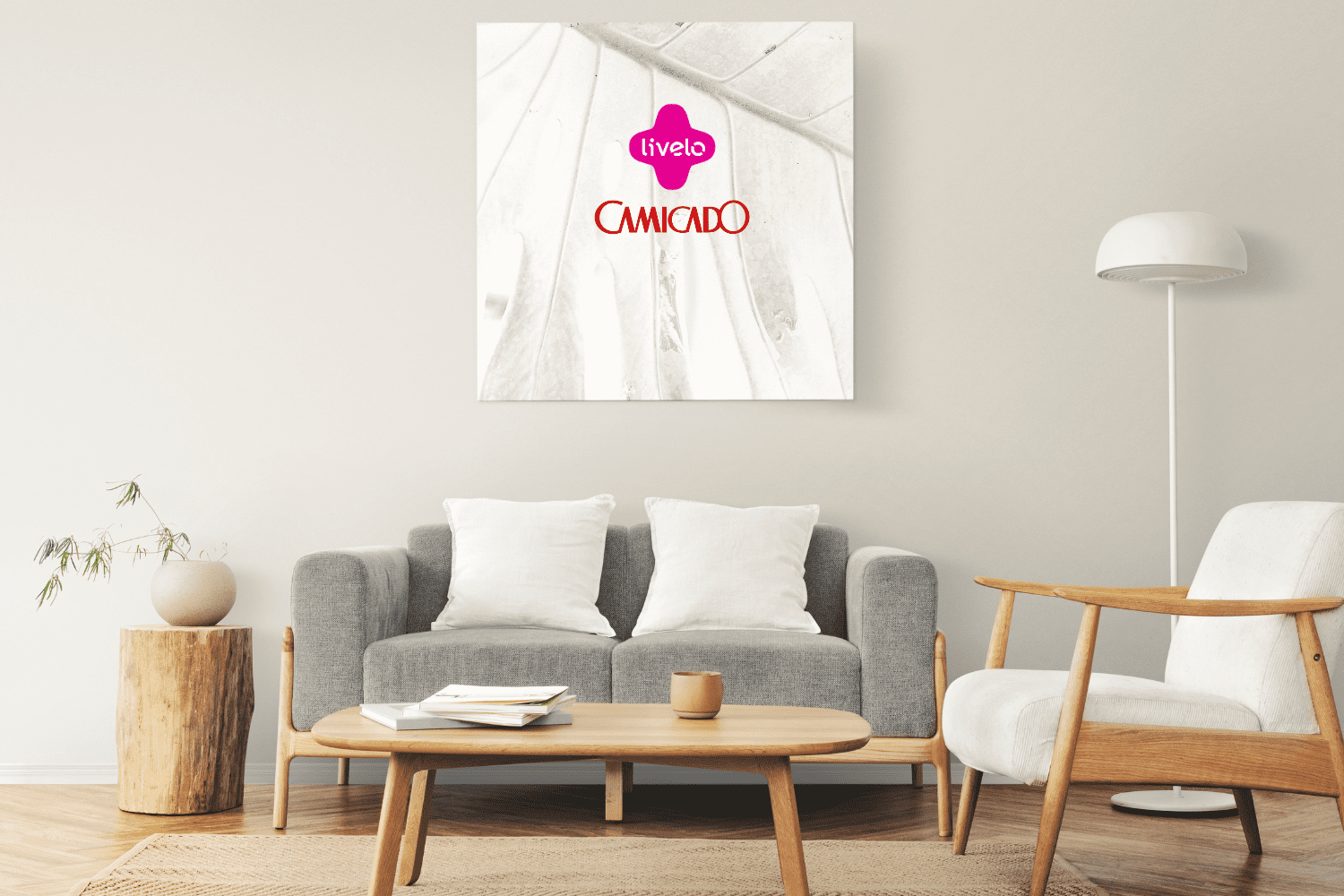 Sala de estar decorada com sofá cinza, almofadas brancas e mesa de centro de madeira. Uma luminária de chão branca e uma poltrona com estrutura de madeira complementam o ambiente. Na parede, um quadro com a logomarca da Livelo e da Camicado.