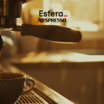 Café fresco sendo preparado em uma máquina de café, com uma xícara de porcelana cheia de café quente. No topo da imagem estão as logos da Esfera e Nespresso, a imagem transmite uma sensação de aconchego e prazer matinal.
