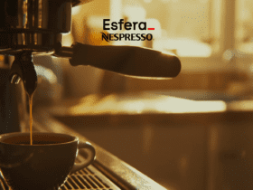 Café fresco sendo preparado em uma máquina de café, com uma xícara de porcelana cheia de café quente. No topo da imagem estão as logos da Esfera e Nespresso, a imagem transmite uma sensação de aconchego e prazer matinal.