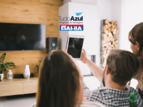 Duas pessoas estão sentadas em um sofá, assistindo a algo em um tablet que uma delas segura, enquanto uma TV grande está montada na parede à frente. No canto superior direito da imagem, estão as logos do programa de vantagens "TudoAzul" e "Casas Bahia".