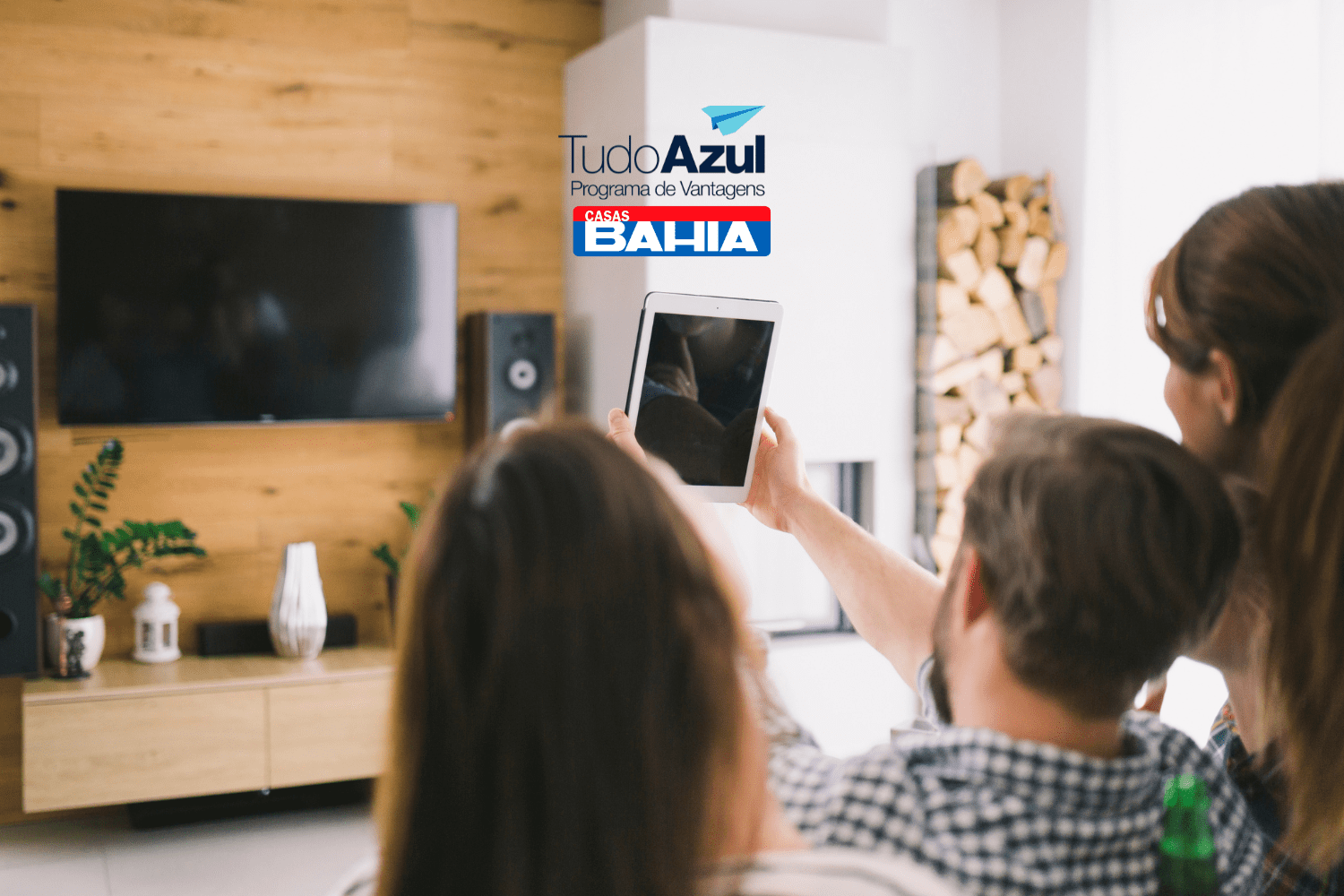 Duas pessoas estão sentadas em um sofá, assistindo a algo em um tablet que uma delas segura, enquanto uma TV grande está montada na parede à frente. No canto superior direito da imagem, estão as logos do programa de vantagens "TudoAzul" e "Casas Bahia".