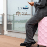 Pessoa encostada em uma mala rosa, segurando um celular, com uma varanda ao fundo. No canto superior esquerdo da imagem, estão as logos do programa de vantagens "TudoAzul" e "Portal das Malas".